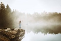 Жінка стояла в білій сукні на скелі, дивлячись на озеро в туманний день — стокове фото