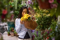 Menina asiática bonita comprando flores na loja de flores enquanto carrega uma cesta de vime com flores amarelas. — Fotografia de Stock