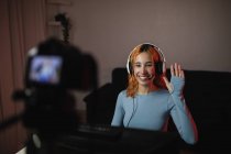 Улыбающаяся женщина-геймер в наушниках машет рукой во время записи видео на профессиональную камеру для блога социальных сетей — стоковое фото
