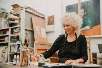 Artista femenina enfocada sentada a la mesa y pintando con acuarelas sobre papel mientras trabaja en un taller creativo - foto de stock