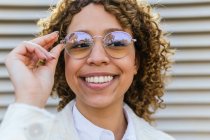 Delighted young African American female with curly hair touching trendy sunglasses and looking away in city — Stock Photo