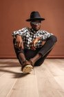 Young cool African American male model in checkered shirt and hat looking at camera while sitting on brown background — Stock Photo