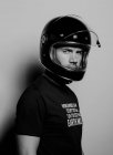 Вид збоку на впевненого брутального дорослого чоловіка в шоломі на чорному мотоциклі, який дивиться на камеру, стоячи на фоні студії — стокове фото