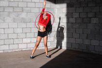 Jeune femme tatouée en vêtements de sport tourbillonnant hula hoop tout en dansant contre des murs de briques avec des ombres et regardant vers l'avant dans la lumière du soleil — Photo de stock