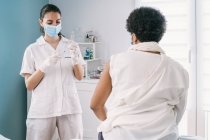 Doctora en guantes de látex y protector facial llenando jeringa de frasco con vacuna preparándose para vacunar a una mujer afroamericana madura irreconocible en clínica durante el brote de coronavirus - foto de stock