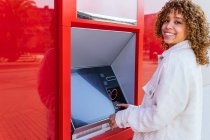 Vue latérale d'une femme afro-américaine souriante utilisant un terminal ATM et retirant de l'argent tout en se tenant debout dans la rue de la ville en regardant la caméra — Photo de stock