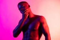 Serious young African American male athlete with naked torso looking at camera and touching face on pink background in neon studio — Stock Photo