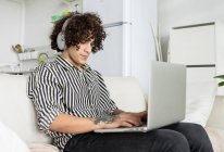Jovem hipster macho com fones de ouvido navegando na internet no netbook enquanto descansa no sofá no quarto da casa — Fotografia de Stock