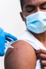 Cultivé spécialiste féminin méconnaissable en uniforme de protection, gants en latex et masque facial vaccinant l'homme afro-américain patient en clinique pendant l'épidémie de coronavirus — Photo de stock