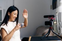 Smiling ethnic female blogger with cup of hot drink showing hello gesture while recording video on photo camera at home — Stock Photo