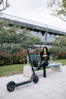 Сосредоточенная женщина-предприниматель сидит на скамейке рядом с современным электроскутером и записывает план в блокнот во время работы в удаленном городском парке — стоковое фото