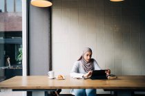 Content Muslim female in hijab and talking on video chat via tablet while sitting at table in cafe — Stock Photo