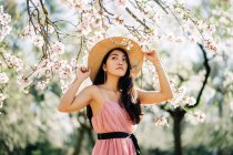 Peaceful ethnic female in straw hat and dress standing under blooming fragrant flowers on tree branches in orchard looking away — Stock Photo