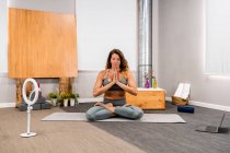 Ganzkörper einer konzentrierten jungen Frau in aktiver Kleidung, die in Lotus-Pose mit geschlossenen Augen und Gebetshänden sitzt und während einer Yoga-Sitzung zu Hause in der Nähe eines Smartphones meditiert, das auf einem Stativ platziert ist — Stockfoto