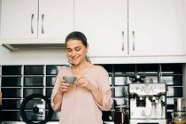 Весёлая женщина средних лет с кружкой горячего напитка, стоящей у кухонного стола с белыми шкафами и современной кофеваркой дома — стоковое фото