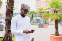 Trendy African American male standing on street with palm trees and messaging on social media via mobile phone — Stock Photo