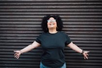 Content adult overweight female in eyewear and open arms curly hair against ribbed wall in daytime — Stock Photo