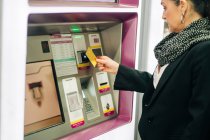 Побочный обзор серьезной женщины, стоящей рядом с банкоматом с картой при покупке билета на поезд, стоя в современном терминале — стоковое фото
