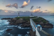 Вражаючі краєвиди моста, що ведуть до скелястого острова, вкритого зеленою травою з маяком, поміщеним у хвилястий океан у Фаро - Ілла - Панча (Галісія, Іспанія) в сутінках. — стокове фото