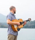 Вид збоку бородатий хлопець в повсякденному одязі, що стоїть з гітарою на дерев'яному пірсі біля річки з горами на фоні під хмарним сірим небом вдень — стокове фото