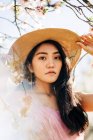 From below peaceful thoughtful ethnic female in straw hat and dress standing under blooming fragrant flowers on tree branches in orchard looking at camera — Stock Photo