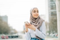 Joven mujer étnica en hijab de pie en la ciudad y mensajería en el teléfono móvil - foto de stock