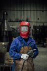 Anonymer Mechaniker mit Schutzhelm und Schürze auf blauem Overall mit Schweißschlauch in leichter Werkstatt in der Nähe von Metallkonstruktionen — Stockfoto