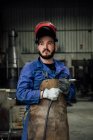 Mechaniker mit Schutzhelm und Schürze auf blauem Overall mit Schweißschlauch in leichter Werkstatt in der Nähe von Metallkonstruktionen — Stockfoto
