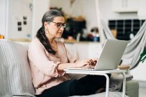 Side view full body of concentrated middle aged female in eyeglasses typing on netbook while sitting on sofa in room with kitchen on blurred background — Stock Photo