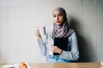 Portrait of Muslim female freelancer in headscarf standing at table with cup of beverage and tablet while thinking about project and looking at camera — Stock Photo