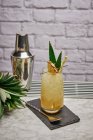 Gelber Cocktail im Glas garniert mit Ananasstück und grünen Blättern mit Papierstroh auf Schieferuntersetzer mit Barlöffel gelegt — Stockfoto