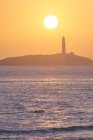 Cenário pitoresco de ondulação de água da costa de lavagem do oceano com farol de elevação colocado em Faro de Trafalgar em Cádiz, na Espanha, sob o céu laranja brilhante ao nascer do sol — Fotografia de Stock