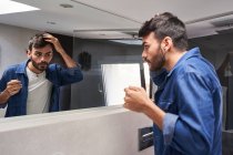 Side view of concentrated young bearded ethnic guy in stylish clothes touching hair while looking in mirror in modern bathroom — Stock Photo