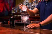 Camarero irreconocible poniendo un gran cubo de hielo en el vaso mientras prepara un cóctel gin tonic en el bar - foto de stock
