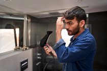 Side view of self assured young bearded Hispanic guy in denim shirt using hairdryer while adjusting hair in bathroom — Stock Photo