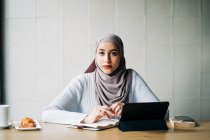 Zufriedene Muslimin im Hidschab, die mit ihrem Tablet arbeitet und im Café am Tisch sitzt, während sie in die Kamera schaut — Stockfoto