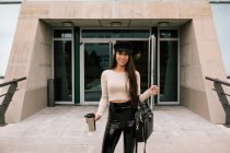 Young happy trendy female entrepreneur standing near entrance of contemporary building holding take away cup in city and looking at camera — Stock Photo