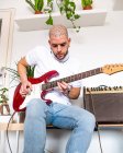 From below of concentrated musician in casual clothes sitting with electric guitar in light room with white walls in daytime — Stock Photo