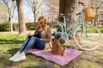 Konzentrierte junge Frau macht sich Notizen in Notizbuch auf kariertem Stoff mit Rucksack in der Nähe von Fahrrad im Park — Stockfoto