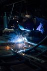 Lavoratore senza volto in guanti e dettagli metallici uniformi di saldatura sul tavolo vicino alle costruzioni in fabbrica — Foto stock