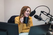 Mulher otimista sentada à mesa com computadores e bebendo bebida quente enquanto fala com o microfone durante o trabalho na estação de rádio moderna — Fotografia de Stock