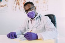 African American doctor in eyeglasses working while writing in patient file at table in hospital — Stock Photo