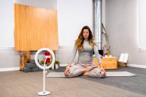 Corps entier de la jeune femme concentrée en vêtements de sport assis dans la pose Lotus avec les yeux fermés faire de la méditation pendant la séance de yoga à la maison près du smartphone placé sur le trépied — Photo de stock