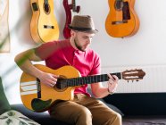 Пенсионер в повседневной одежде и шляпе играет на гитаре в светлой комнате дома в дневное время — стоковое фото