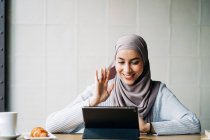 Hembra musulmana en hiyab saludando con la mano y hablando en el chat de vídeo a través de la tableta mientras está sentado en la mesa en la cafetería - foto de stock