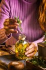 Crop faceless female in purple sweater putting herbs twigs with green leaves in essential oil glass bottle near scissors and rope with small chest on cloth at wooden table — Stock Photo