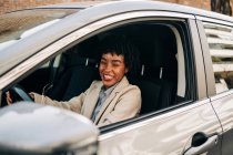 Vista lateral do alegre piloto feminino afro-americano na roupa da moda sorrindo enquanto dirige o automóvel moderno na rua — Fotografia de Stock