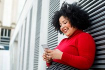 Side view of plump smiling female in bright wear text messaging on cellphone while leaning on ribbed wall in town — Stock Photo