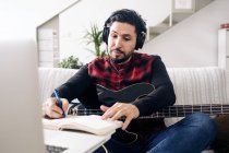 Erwachsener männlicher Gitarrist mit Kopfhörer und E-Gitarre macht sich Notizen im Notizbuch, während er zu Hause auf dem Sofa Musik gegen Netbook komponiert — Stockfoto
