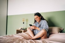 Позитивная латиноамериканка средних лет с длинными темными волосами в повседневной одежде, улыбающаяся, сидя дома на кровати в телефоне — стоковое фото